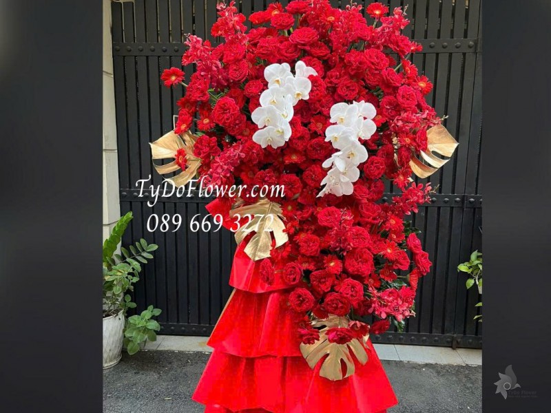 KCM0126027 KỆ HOA CHÚC MỪNG KỶ NIỆM NGÀY THÀNH LẬP Thiết kế hoa đồng tiền đỏ Red Ohara Rose, Lan Hồ Điệp, Lá Trầu Bà Nam Mỹ, Lan Mokara, Đồng Tiền Đỏ