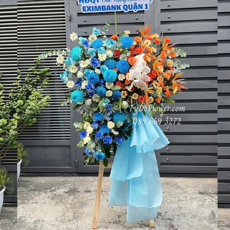 KCM0625490 KỆ HOA CHÚC MỪNG KHAI TRƯƠNG Thiết kế gam màu cam xanh dương, chủ đạo hoa Cúc Mẫu Đơn Xanh Dương, Cam Sprit Roses, Thiên Điểu