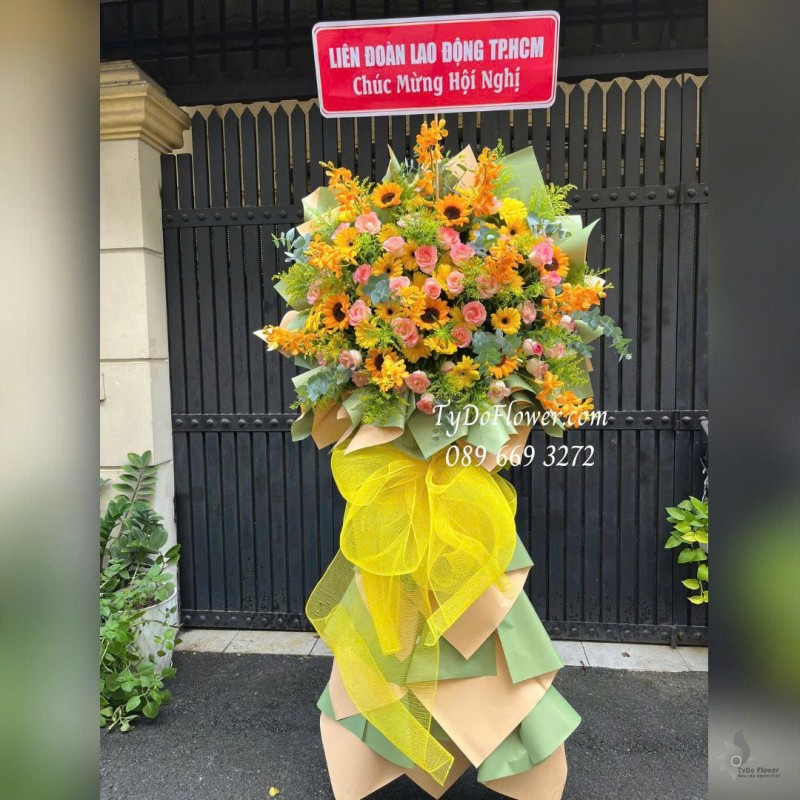 KCM1025849 KỆ HOA CHÚC MỪNG HỘI NGHỊ Thiết kế hoa hồng Lạc Thần, lan Mokara, Hướng Dương, Đồng Tiền Vàng trên nền giấy mỹ thuật cao cấp