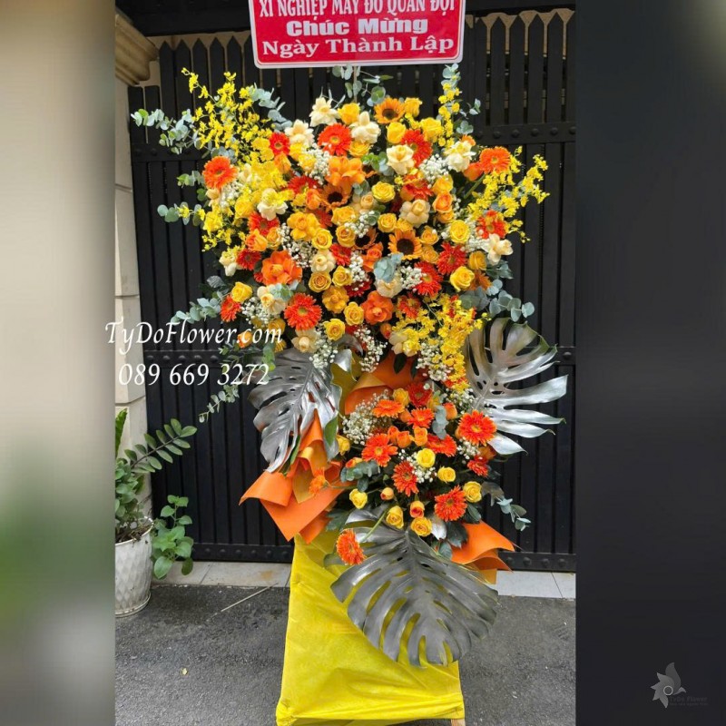 KCM0126028 KỆ HOA CHÚC MỪNG NGÀY THÀNH LẬP Thiết kế tông màu vàng cam, hoa hồng, hoa đồng tiền, lan vũ nữ, hoa hướng dương, lá trầu bà nam mỹ